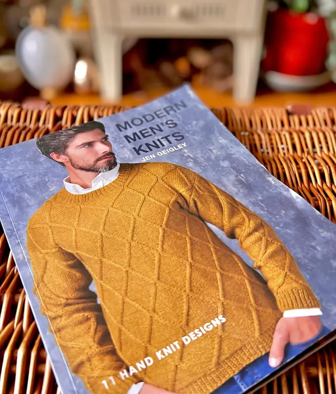 The book Modern Men's Knits by Jen Geigley on a wicker basket, with a man in a mustard textured jumper on the cover