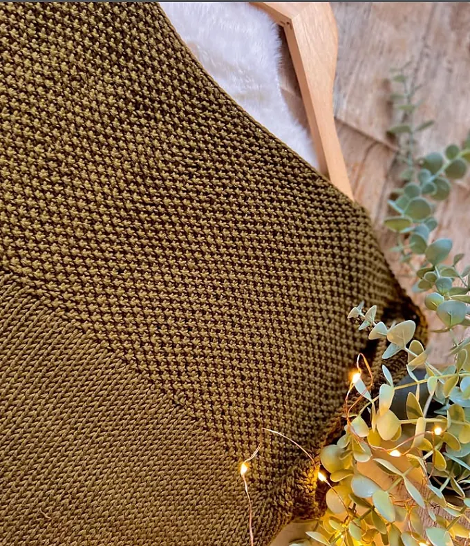An olive moss stitch top laid on a wooden hanger with eucalyptus and fairy lights