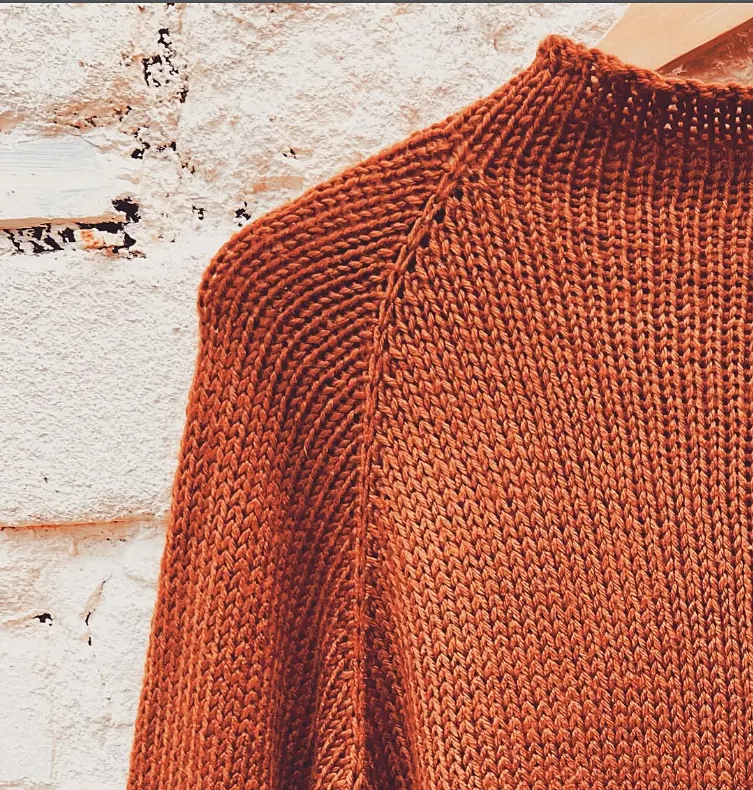 Close up of the raglan shoulder of a burnt orange knitted jumper on a hanger against a white brick wall