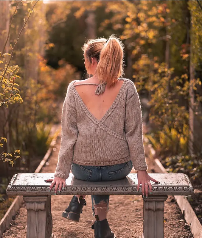 Emma sitting on a stone bench in an autumn garden, wearing her hand knitted open back jumper in oatmeal