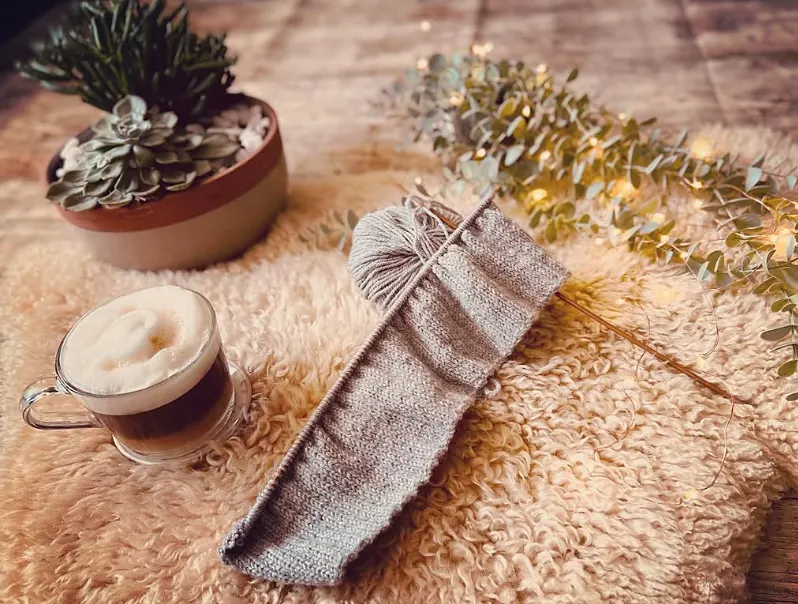A grey knitting project on wooden needles, on a sheepskin rug beside a coffee, a succulent and eucalyptus with fairy lights